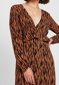 Donna che indossa un vestito a maniche lunghe con strisce di tigre marroni e nere, scollatura a V profonda e una collana con pendente circolare argentato, mano sulla vita.