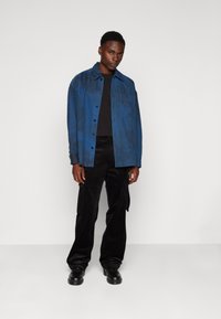 Blå skjorte med knepping og brystlomme, brukt over en svart t-skjorte. Svart kordfløyels cargo-bukser og svarte sko, stående mot en lys bakgrunn.