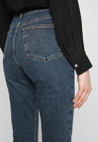 Mörka denimjeans med en figurnära design, som har bakfickor, subtila sömnadsdetaljer och en medelhög midja. Slät textur.