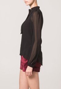 Schwarze Bluse mit transparenten langen Ärmeln, kragenförmigem Ausschnitt und leicht tailliertem Saum. Kombiniert mit bordeauxroten Shorts. Glatte Textur.