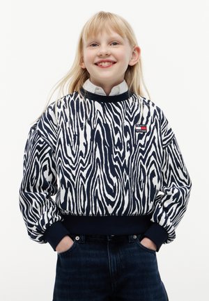 Jeune fille souriante aux cheveux blonds portant un pull à motifs noir et blanc et un jean foncé, les mains dans les poches, debout devant un fond uni.