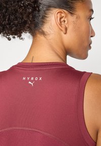 Haut de sport sans manches bordeaux avec une texture lisse, arborant le logo "HYROX" et l'emblème Puma situés à l'arrière près de l'encolure.