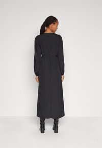 Rochie maxi neagră cu mâneci lungi, talie ajustată și o textură netedă. Prezintă un detaliu la gâtul din spate și cizme negre până la gleznă.