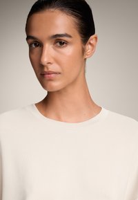 Világos bézs, bordázott felső, kerek nyakkal. Simább textúra látható. Közeli felvétel a felső testről és az arcról, lágy, semleges háttér előtt.