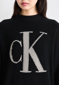 Črn pleteni pulover s velikim bež "CK" logotipom v teksturiranem dizajnu. Rebrast ovratnik in sproščen kroj.