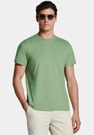 Homme portant des lunettes de soleil rondes, t-shirt manches courtes vert clair, pantalon beige, debout avec une main dans la poche sur fond blanc.