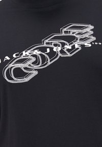 Juodas marškinėlis su dideliu, baltu grafiniu dizainu, kuriame išryškintos 3D blokinės raidės ir aiškiai matomas užrašas "JACK & JONES 1999".