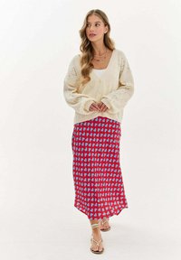 Krémový pletený kardigán s výstrihom do V a vzorom srdcí, skombinovaný s červenou sukňou s modrými abstraktnými tvarmi. Celkový vzhľad dopĺňajú sandále na šnúrky.