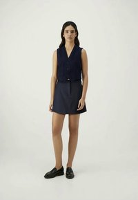 Colete sem mangas azul-marinho com decote em V e fecho de botão, combinado com uma mini-saia azul-marinho a condizer e sapatos pretos. Design simples e limpo.