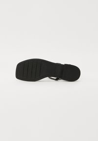 Musta sandaalin pohja, jossa on teksturoitu kumipinta. Ominaisuudet ovat suorakaiteen muoto ja urat pidon lisäämiseksi. Minimalistinen muotoilu, ilman näkyvää metalliosaa.