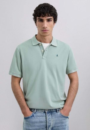 Camisa polo de algodón en verde claro con mangas cortas, tapeta de tres botones y pequeño logo de calavera verde oscuro en el lado izquierdo. Combinada con jeans de mezclilla azul.