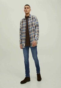 Karierter Hemd in Blau und Creme, mit Knöpfen und zwei Fronttaschen, über einem braunen Shirt getragen. Kombiniert mit blauen Jeans und braunen Stiefeln.