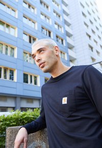 Cămașă neagră din bumbac cu mânecă lungă, dotată cu un buzunar pe piept, având un patch cu logo galben. Croiala este structurată, fiind prezentată pe un fundal urban estompat.