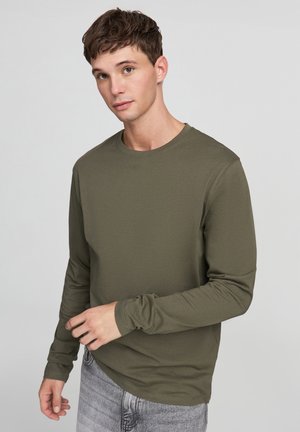 T-shirt a maniche lunghe verde oliva con scollo rotondo, texture morbida, maniche attillate e cuciture sottili abbinato a jeans grigi chiari.