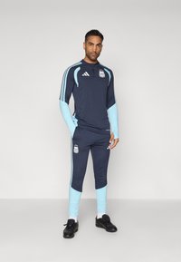 Άνδρας ντυμένος με ναυτικό μπλε και ανοιχτό μπλε προπονητική στολή της Εθνικής Αργεντινής, με λογότυπα της Adidas σε φανέλα και παντελόνια, φορώντας μαύρα παπούτσια.