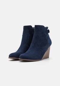 Bottines en suède bleu marine avec un talon compensé, un bout rond, une fermeture éclair sur le côté et une sangle décorative avec un détail en métal. Semelle plate marron.