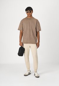 Braunes oversized T-Shirt mit Rundhalsausschnitt und sichtbaren Nähten, kombiniert mit hellbeigen, schmal zulaufenden Jogginghosen und einer schwarzen Handtasche.