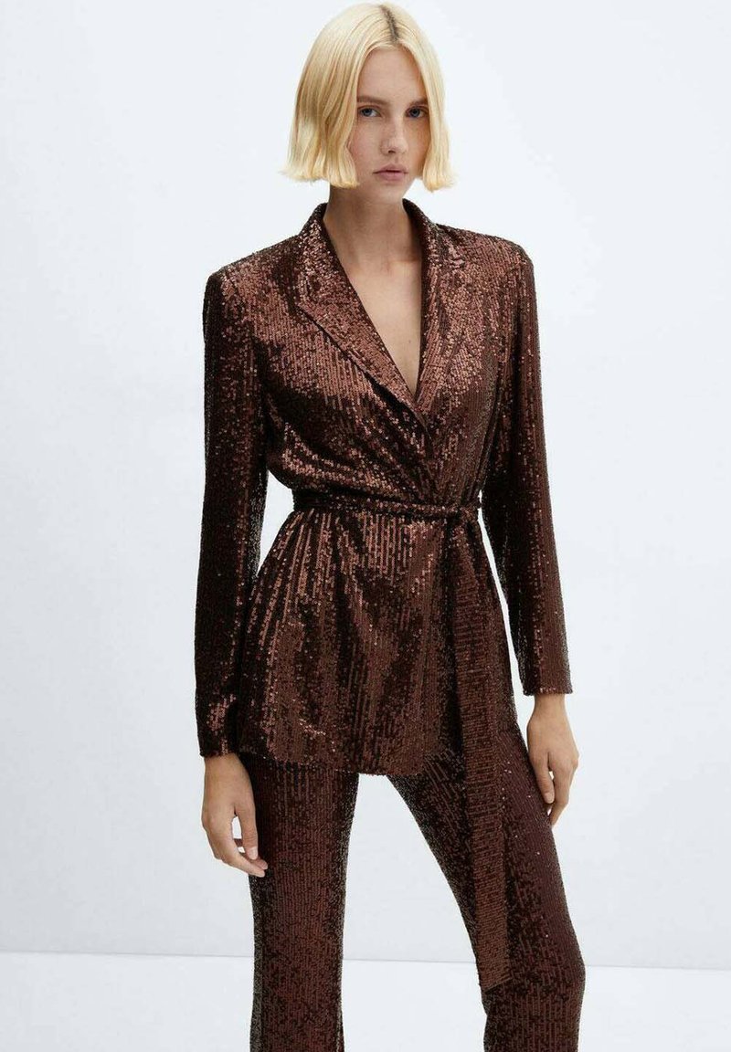 Donna con capelli biondi corti che indossa un blazer marrone con paillettes e pantaloni abbinati, in piedi davanti a uno sfondo bianco semplice.