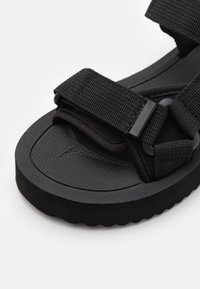 Mustat sandaalit, joissa on teksturoitu kumipohja, säädettävät kangasnauhat ja pehmustettu pohja mukavuutta varten. Littee muotoilu saumattomalla viimeistelyllä.