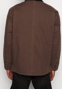 Person wearing a brown jacket with black collar, standing with hands relaxed by sides, shown from the back against a plain background.