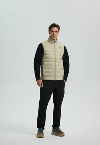 Uomo in piedi che indossa un gilet trapuntato beige sopra una maglia nera a maniche lunghe, pantaloni cargo neri e scarpe da trekking marroni con lacci blu.