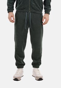 Pantaloni jogger in corduroy verde scuro con vita elasticizzata, cordoncino e gambe affusolate, abbinati a scarpe da ginnastica di colore chiaro.