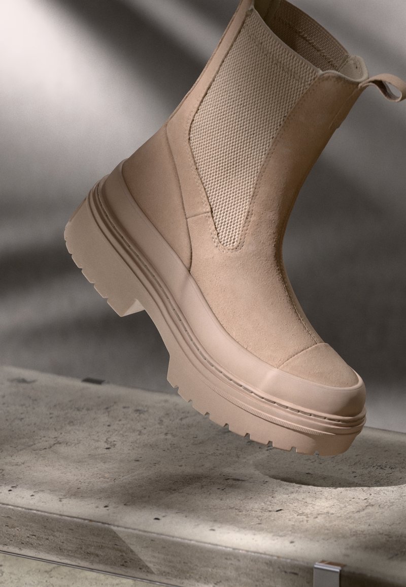 Botte Chelsea en daim beige avec semelle en caoutchouc épaisse et panneau latéral élastique, suspendue au-dessus d'une surface en béton avec un éclairage doux.