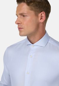 Himmelblaues Hemd mit Knopfleiste aus einem glatten Material, einem spitzen Kragen und einem halbdurchsichtigen Stoff, der zwei Knöpfe am Kragen hat.