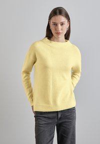Žlutý pletený svetr s vysokým výstřihem, který má na jedné straně ribbed texturu a na druhé hladký povrch. V kombinaci s tmavými džíny.