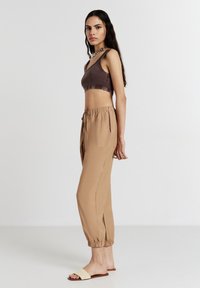Top corto marrone abbinato a pantaloni jogger beige, morbidi e con elastico alle caviglie. Modella in sandali scivolati beige.