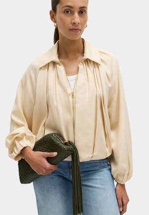 Kvinde iført en cremefarvet bluse med lynlås og puffede ærmer samt lyseblå jeans, der holder en mørkegrøn vævet clutch med kvaster.