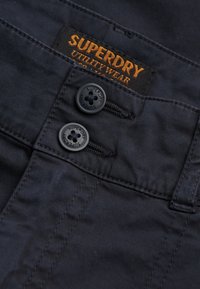 Pantalon bleu marine foncé à texture lisse, avec deux boutons noirs et une étiquette orange portant l'inscription « Superdry Utility Wear ».