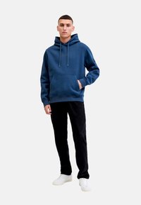 Sudadera azul marino con cordones, bolsillo frontal y detalle de logotipo. Combinada con pantalones de pana negros y zapatillas blancas.