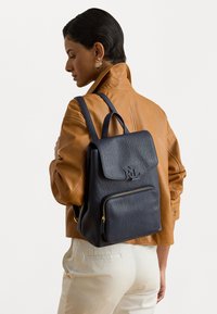 Σκούρο μπλε δερμάτινος σακίδιο με καπάκι στην κορυφή, μπροστινή τσέπη με φερμουάρ και χρυσές λεπτομέρειες. Υφή που προσομοιώνει και διπλοί ιμάντες ώμου.