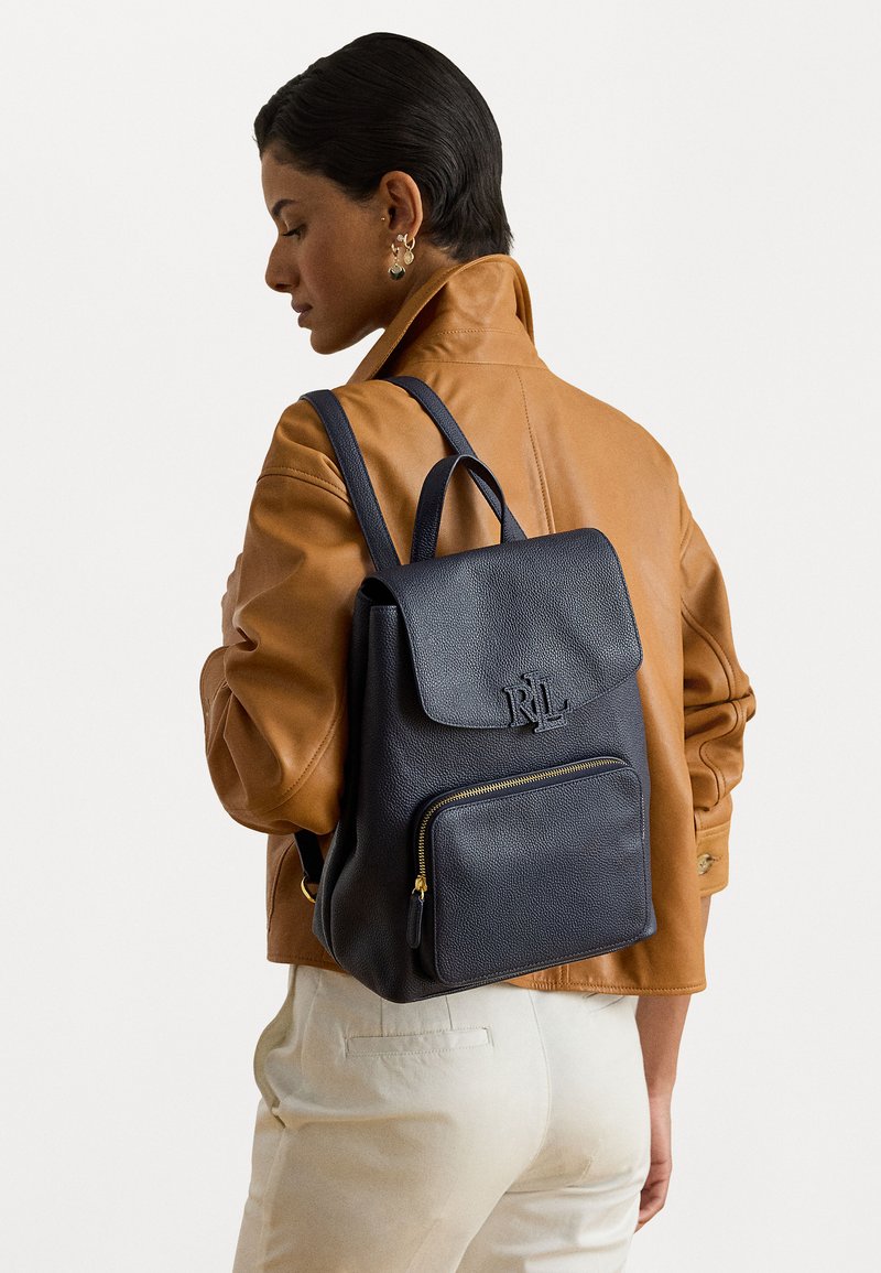 Σκούρο μπλε δερμάτινος σακίδιο με καπάκι στην κορυφή, μπροστινή τσέπη με φερμουάρ και χρυσές λεπτομέρειες. Υφή που προσομοιώνει και διπλοί ιμάντες ώμου.