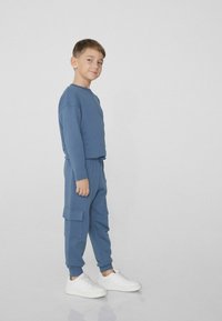 Modré mikina a kalhoty s cargo stylem, vyrobené z měkké látky, s elastickými manžetami, šňůrkovým pasem a bočními kapsami, doplněné bílými teniskami.