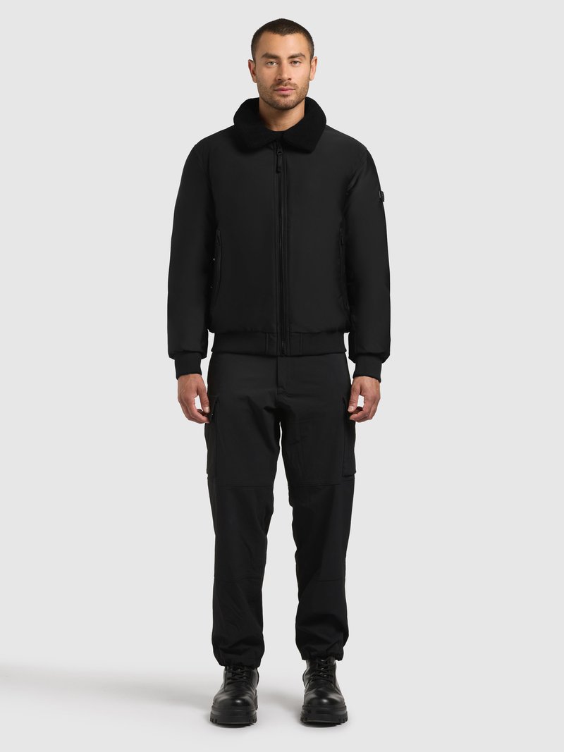 Melnā bumbvedēja jaka ar mīkstu apkakli, rāvējslēdzēja priekšpusi un ribām piedurknēm, kopā ar melnām kravas biksēm un izturīgiem melniem zābakiem.