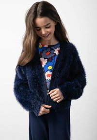 Granatowa kurtka z sztucznego futra z zamkiem błyskawicznym, długimi rękawami i miękką teksturą, noszona na kolorowej bluzce w kwiatowy wzór.
