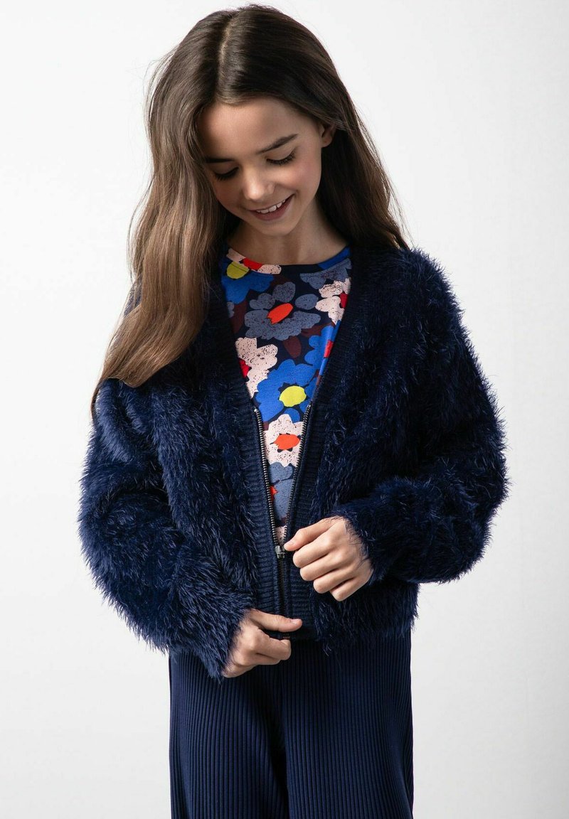 Granatowa kurtka z sztucznego futra z zamkiem błyskawicznym, długimi rękawami i miękką teksturą, noszona na kolorowej bluzce w kwiatowy wzór.