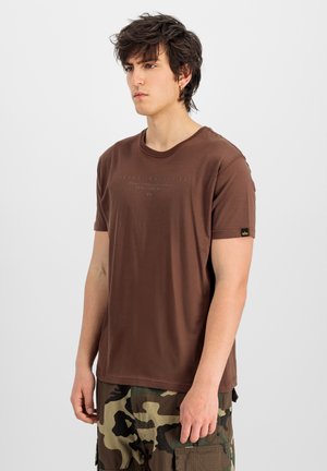 Junger Mann mit zerzausten dunklen Haaren, trägt ein braunes Alpha Industries T-Shirt und Camouflage-Cargo-Shorts, schaut leicht nach links vor einem einfarbigen Hintergrund.