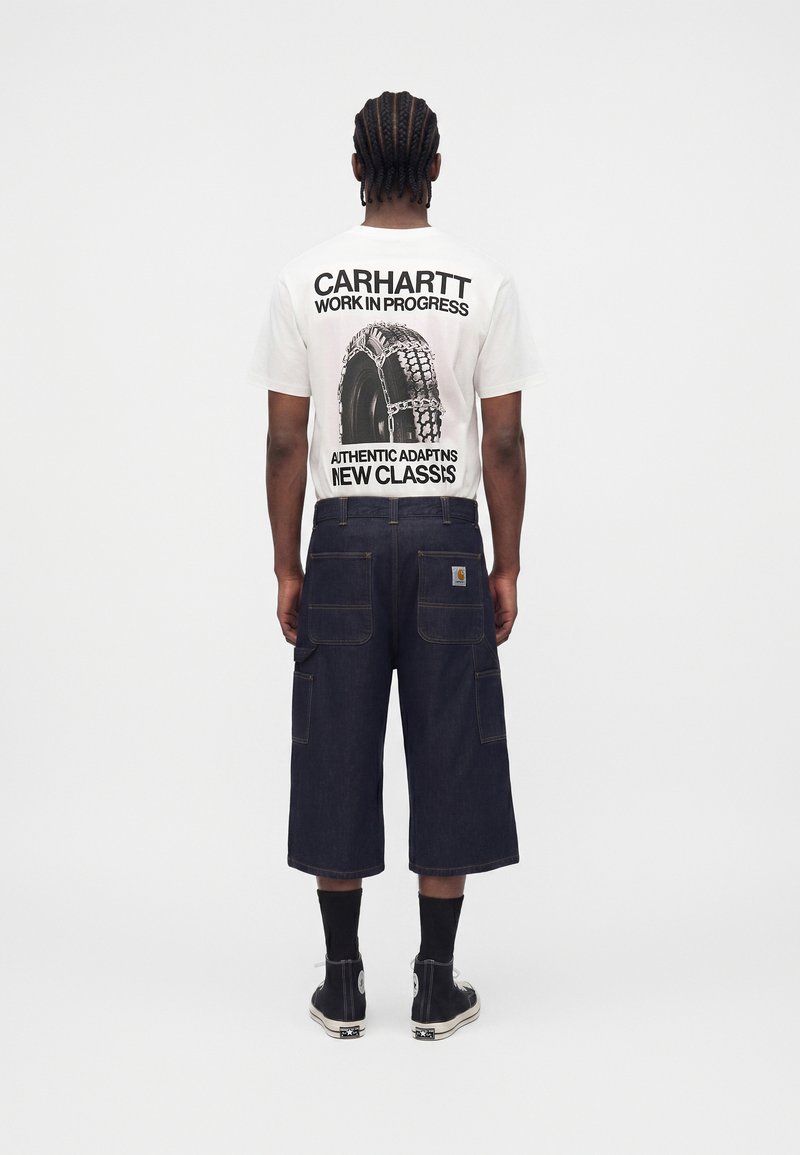 Mand med fletninger iført hvid Carhartt grafisk t-shirt, mørke vidde cargo shorts, sorte sokker og sorte høje sneakers, med ryggen mod.