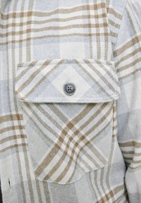 Camisa de franela a cuadros en gris y beige con un bolsillo en el pecho y un solo botón gris. Presenta una textura suave y un patrón de cuadros clásico.