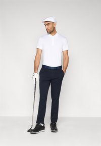 Άσπρο πόλο μπλουζάκι γκολφ, μπλε σκούρο παντελόνι, λευκό καπέλο, μαύρα παπούτσια γκολφ, κρατώντας μπαστούνι γκολφ, φοράει λευκό γάντι, κανονική εφαρμογή χωρίς σχέδια.