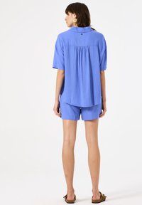 Blaues Leinenhemd mit kurzen Ärmeln, mit einem Faltendetail am Rücken, kombiniert mit passenden blauen Shorts. Das Model trägt braune Sandalen.