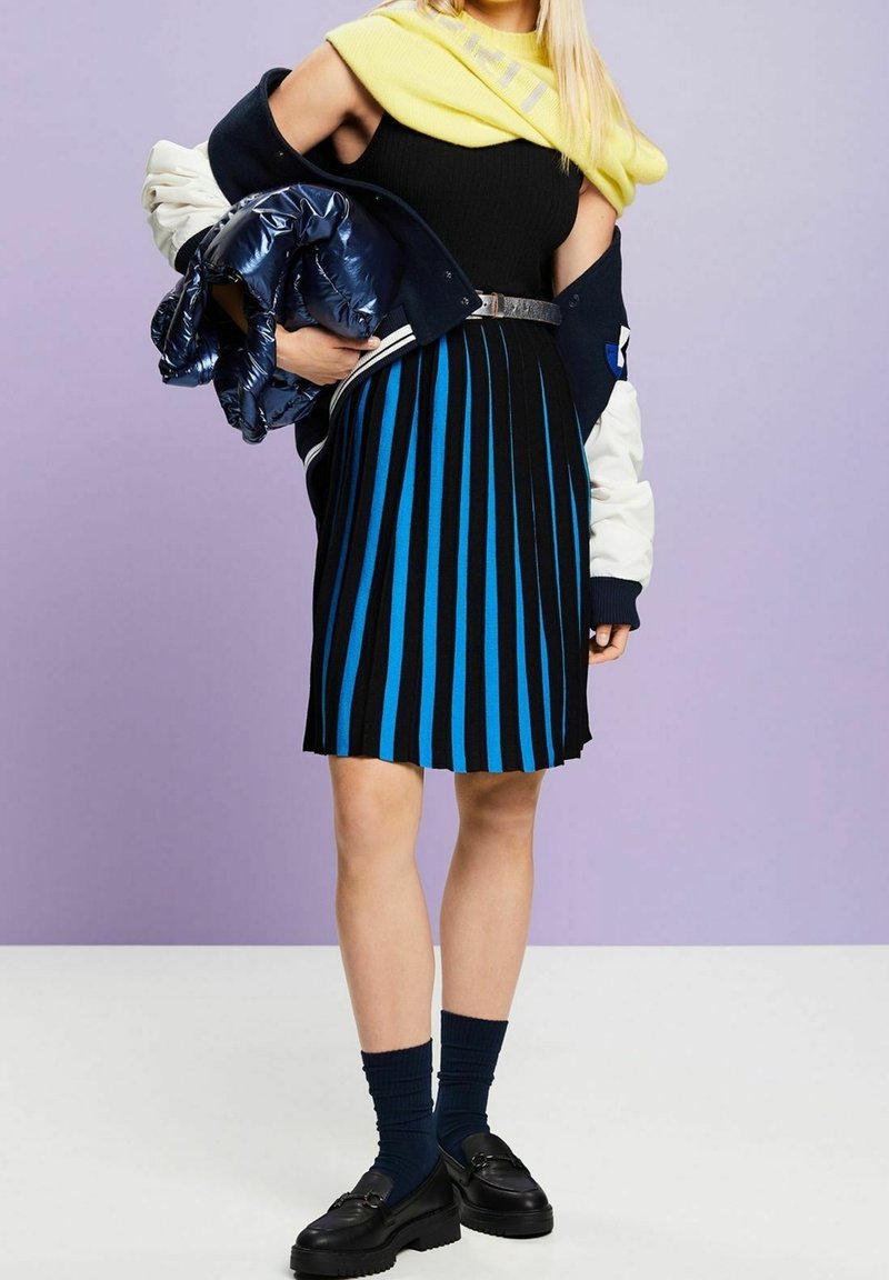 Femme portant une jupe rayée noire et bleue, un haut noir, des chaussettes bleu marine, des chaussures noires, avec une écharpe jaune et tenant une veste bleu marine sur un bras.