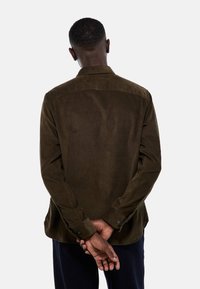 Chemise en velours côtelé marron à manches longues, avec un col évasé et des détails au niveau du dos. Poignets à boutons, texture lisse et coupe décontractée.