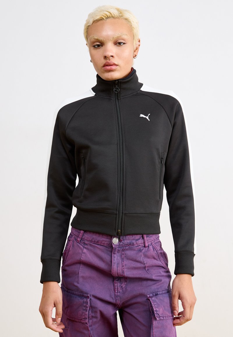 Black zip-up jacket with a high collar, features white shoulder stripes and a small white logo on the chest, made from a smooth fabric.