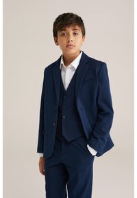 Marinblå kostym med blazer, väst och byxor, med en vit skjorta under. Slät tyg med strukturerad design och knappar.