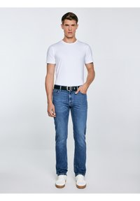 Vīrietis stāv, valkājot vienkāršu baltu T-kreklu, zilas džinsas ar melnu jostu un baltas sporta kurpes, pret vienkāršu gaišu fonu.