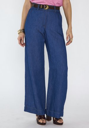 Femme portant un pantalon en jean bleu à taille haute et jambes larges, ceinture marron, top rose, bracelets dorés, bagues et sandales marron à plateforme et bout ouvert.